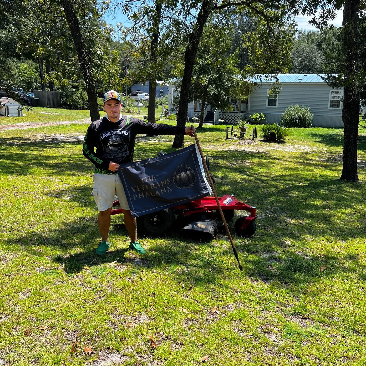 A Lawn Rangers crew member holding The Veterans Phalanx Standard in the yard of an elder veteran after donating a lawn cut.